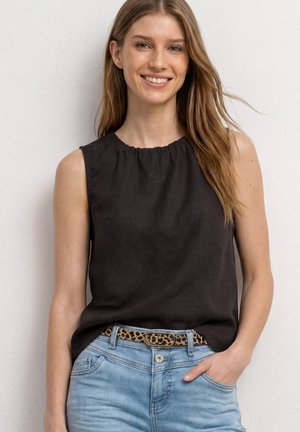 Mujer sonriente con cabello largo castaño claro, viste una camiseta negra sin mangas, jeans azul claro y cinturón con estampado de leopardo, de pie contra una pared blanca.
