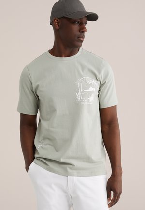 Mann trägt ein hellgrünes T-Shirt mit Grafikmotiv einer Palme und einem Boot, weiße Hose und graue Baseballkappe, steht vor beige Hintergrund.