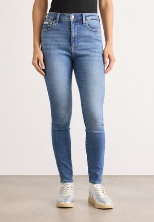 Vaqueros de denim azul de cintura alta, ajustados, con desvanecimiento en los muslos y un bolsillo frontal. Combinados con zapatillas grises claras.