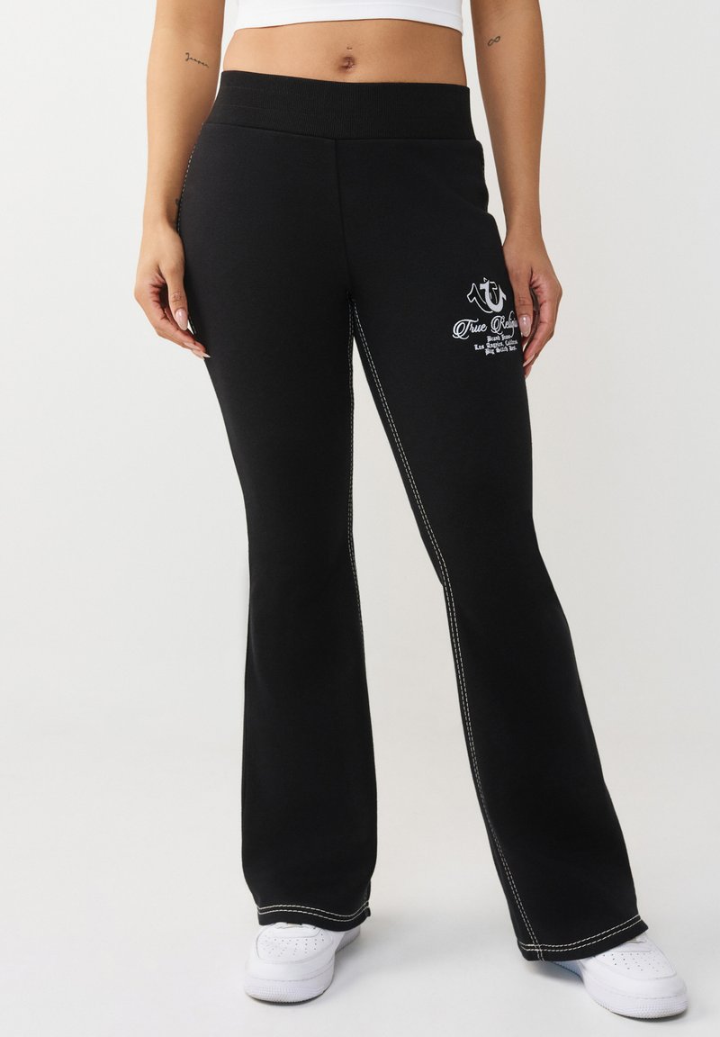 Femme portant un pantalon évasé noir avec des coutures blanches et un logo sur la cuisse gauche, associé à des baskets blanches et un top court blanc.