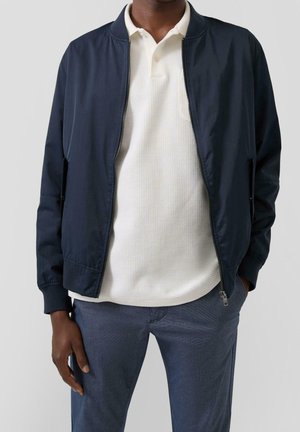 Homme portant une veste bleu marine zippée sur un polo blanc texturé et un pantalon gris, debout avec une main dans la poche.
