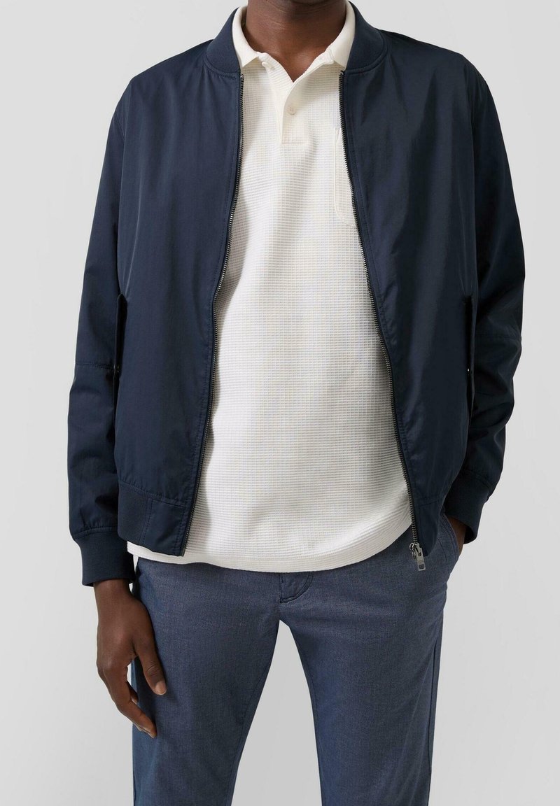 Homme portant une veste bleu marine zippée sur un polo blanc texturé et un pantalon gris, debout avec une main dans la poche.