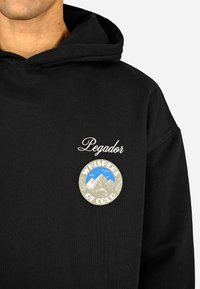 Czarna bluza z kapturem z przednią kieszenią; posiada białe napisy "Pegador" oraz okrągły emblemat z napisem "SZEPTY ZIMNA" i grafiką gór.