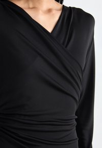 Black wrap dress with long sleeves, featuring a V-neckline and draped fabric, showcasing smooth texture and gathered detailing at the waist.