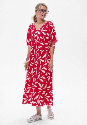 Femme aux cheveux blonds en queue de cheval, portant de grandes lunettes, une robe maxi rouge avec un motif de feuilles blanches, des baskets blanches et des boucles d'oreilles créoles, se tenant debout.