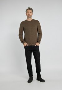 Brązowy sweter z dzianiny o teksturowanym wzorze, zestawiony z czarnymi dżinsami i ciemnymi botkami za kostkę. Model stoi na jednolitym tle.