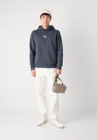Kapuca v navy barvi, bele jeans hlače in bele športne copate. Model drži majhno bež torbico s zgornjim ročajem.