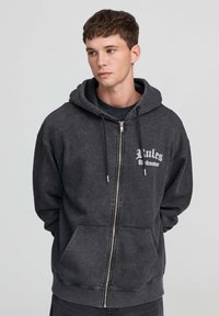 Giovane uomo che indossa una felpa nera con zip sbiadita e tasche frontali, con la scritta bianca "Rules Takedown" sul petto, che guarda di lato.