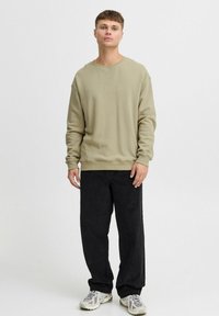 Sudadera caqui con logotipo tonal, cuello redondo y puños de canalé, combinada con pantalones anchos negros y zapatillas beige. Fondo liso.