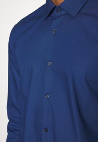 Primo piano di una persona che indossa una camicia a maniche lunghe, blu scuro, con colletto classico.
