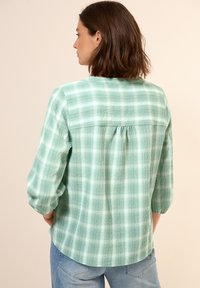 Camicia a quadri verdi in tessuto testurizzato, maniche a tre quarti e dettaglio di arricciatura sul retro, abbinata a un paio di jeans azzurri chiari.