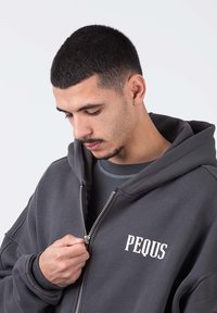 Felpa grigia con zip, grande cappuccio, polsini a coste e tasca frontale. Presenta il testo ricamato in bianco "PEQUS" sull'area sinistra del petto.