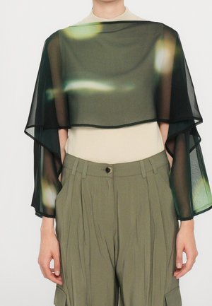 Person trägt einen transparenten schwarzen Poncho mit breiten Ärmeln über einem beigen Top und hoch taillierte olivgrüne plissierte Hose mit Knöpfen vorne.