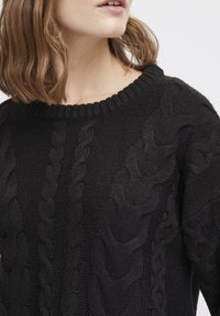 Svart kabelstickad tröja med ribbat rundhalsat och framträdande texturerade mönster fram och på ärmarna. Mjuk, tjockstickad material.