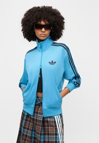 Ανοιχτό μπλε μπουφάν με φερμουάρ, με μαύρες τρεις ρίγες και λογότυπο στο στήθος, συνδυασμένο με καρό καφέ φούστα με μπλε λεπτομέρειες.