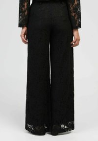 Femme portant un pantalon en dentelle large à taille haute noir et des manches en dentelle assorties, se tenant devant un fond clair uniforme.