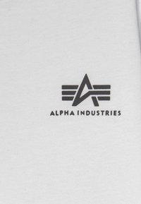 Balts kokvilnas audums ar melnu logo, kurā ir stilizēti burti "A" un trīs horizontālas līnijas virs "ALPHA INDUSTRIES". Mīksta tekstūra.
