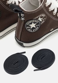 Braune Canvas-Sneaker mit hohem Schaft, weißen Schnürsenkeln, schwarzen Logo-Patches und weißen Gummisohlen, daneben zwei schwarze Schnürsenkel.
