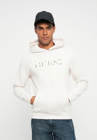 Sudadera blanca con un gran logo en relieve de "GUESS" en el frente. Presenta un bolsillo tipo canguro y capucha ajustable con cordón. Jeans de denim casuales.