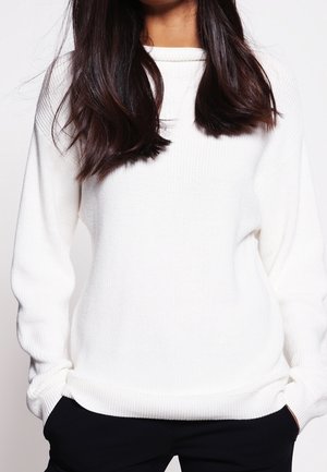 Femme aux longs cheveux foncés portant un pull en maille blanc à manches longues et un pantalon noir, les mains dans les poches, sur un fond uni.