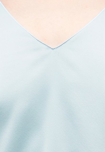 Ελαφρώς γαλάζιο ένδυμα με V λαιμόκοψη πάνω σε γυμνό δέρμα, που δείχνει την απαλή υφή του υφάσματος και τα τακτοποιημένα ράμματα κατά μήκος της λαιμόκοψης.