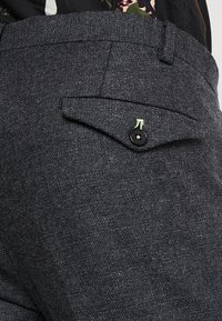Grijze tweed broek met een klepzak met een zwarte knop en neon groene accentstiksels. Textiel met textuur en op maat gemaakt ontwerp.