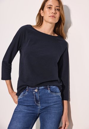 Dunkelblaues langärmliges Oberteil mit weitem Halsausschnitt, kombiniert mit Jeans aus Denim, die über einen Knopfverschluss und dezente Verwaschungen verfügen.