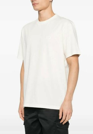 Uomo che indossa una maglietta bianca a maniche corte e pantaloni cargo neri, in piedi davanti a uno sfondo bianco con le mani rilassate lungo i fianchi.