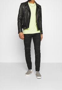 Černá kožená motocyklová bunda, neonově zelené tričko, černé džíny se zipy a kostičkované slip-on boty. Minimalistické doplňky.