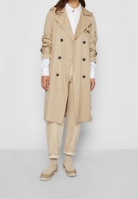 Beige trenchcoat med dobbeltknapdesign, sorte knapper, parret med en hvid skjorte og beige bukser, suppleret med ankelstøvler.
