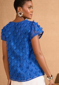 Top blu trasparente con applicazioni floreali, maniche corte a sbuffo e bordi scallop. Accessori in metallo visibili vicino al colletto.