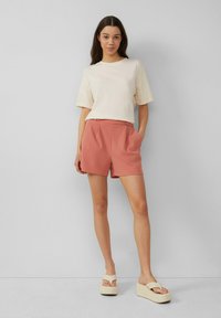 Lichtbeige t-shirt met korte mouwen gecombineerd met roze geplooide shorts. Model draagt crèmekleurige platform sandalen. Soepele texturen, casual ontwerp.