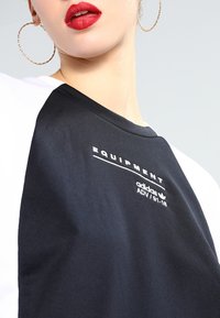 Svart och vit kortärmad skjorta med fet text "EQUIPMENT" och "adidas ADV/91-18" i vitt på svart bakgrund. Slät tygtextur.