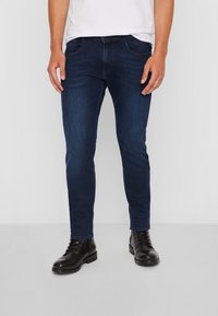 Man som bär mörkblå slim-fit jeans, vit t-shirt och svarta snörkängor av läder, står mot en enkel vit bakgrund.