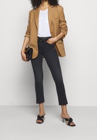 Mujer con blazer camel, camiseta blanca, vaqueros negros cortos con bajos sin rematar, cinturón negro, sandalias negras de tacón y sujetando un bolso negro de mano.