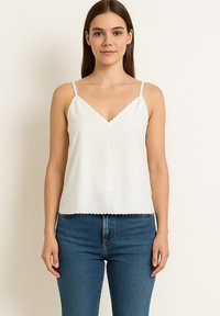 Giovane donna che indossa un top bianco con bordo scalloped e jeans blu, in piedi contro uno sfondo chiaro uniforme.