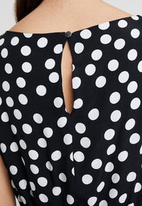 Robe noire à pois blancs, avec une ouverture en forme de goutte d'eau dans le dos fermée par un bouton. Texture douce et coupe décontractée.