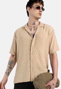 Camisa de manga corta beige con un patrón texturizado, botones en la parte delantera y un corte holgado. El modelo sostiene un artículo acolchado de color oliva.