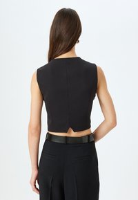 Top court sans manches noir avec un col haut et une petite fente dans le dos, associé à un pantalon taille haute et une ceinture sombre. Texture lisse.