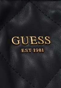 Czarna pikowana skóra z złotym logo „GUESS” i napisem „EST 1981” poniżej, o gładkiej fakturze i przeszyciach w kształcie diamentów.