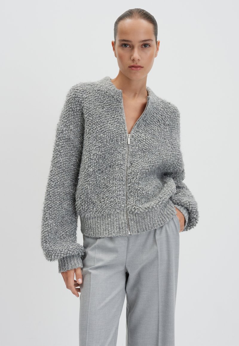 Felpa grigia con zip realizzata in tessuto testurizzato e soffice, con polsini e orlo a coste. Abbinata a pantaloni tagliati grigio chiaro.