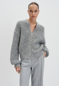 Grauer Zip-Hoodie aus strukturiertem, flauschigem Stoff mit gerippten Bündchen und Saum. Kombiniert mit hellgrauen, taillierten Hosen.