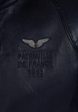 Tissu noir texturé avec broderie "Patrouille de France 1953" et logo d'aile argenté. Le design présente une forme arrondie et des accents contrastants.