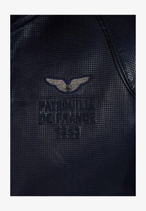 Tissu noir texturé avec broderie "Patrouille de France 1953" et logo d'aile argenté. Le design présente une forme arrondie et des accents contrastants.