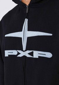 Sudadera con cremallera negra que presenta un gran logo bordado en azul claro con un elemento estrellado y el texto "PXP" en el frente. Textura de tela suave.