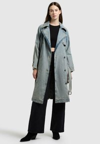 Giovane donna che indossa un trench in denim azzurro chiaro sopra un crop top nero e pantaloni neri a gamba larga, in piedi davanti a uno sfondo neutro.