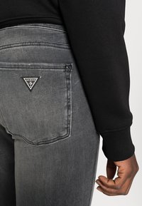 Svart långärmad tröja i kombination med åtsittande grå denimjeans med en triangulär Guess-logotyp på bakfickan. Texturen verkar slät och sliten.