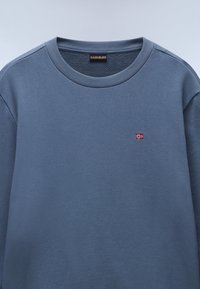 Blå hettegenser med ribbestrikket halsringning og ermer. Har et lite brodert norsk flagg på brystet. Myk tekstur.