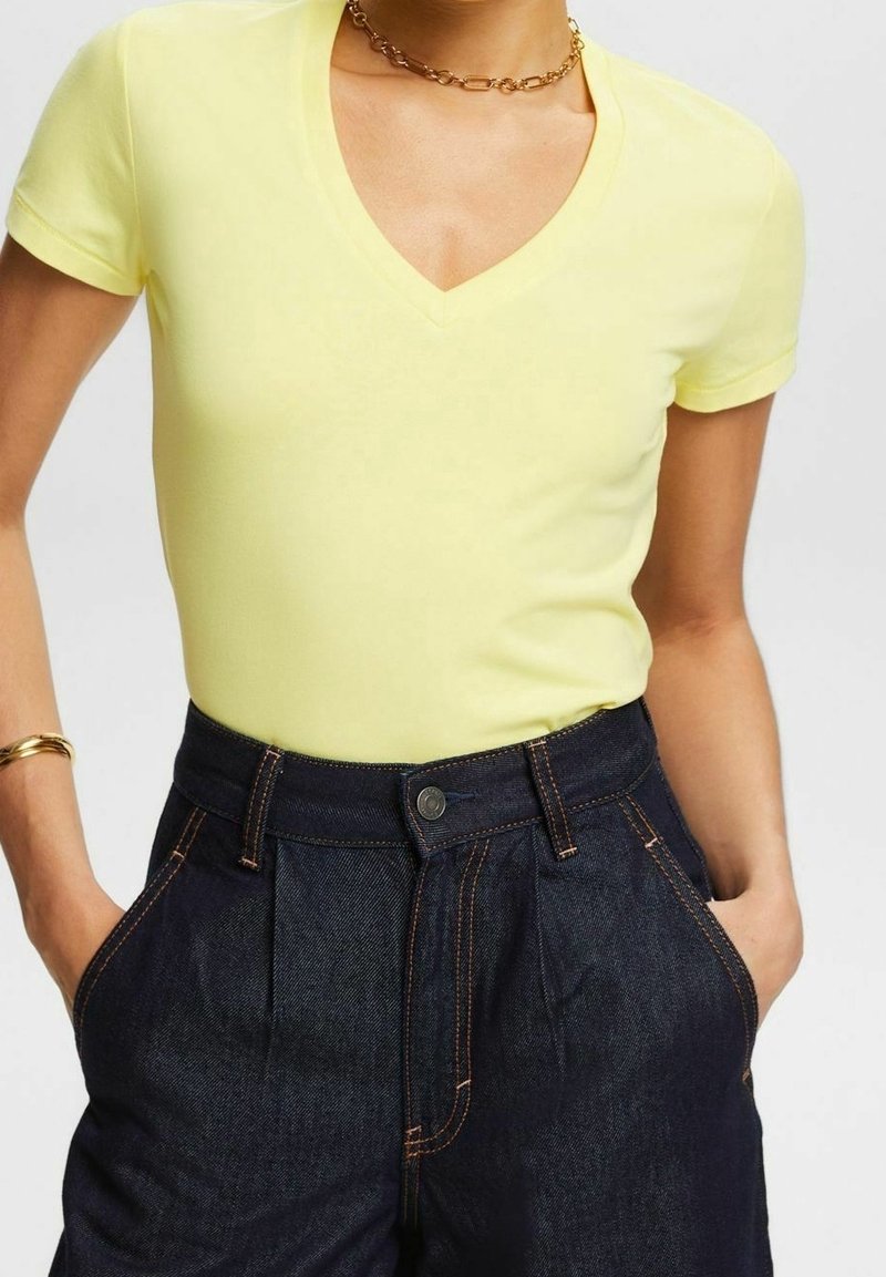 Personne portant un t-shirt jaune à manches courtes en V, rentré dans un jean taille haute bleu foncé, les mains dans les poches et un bracelet doré ainsi qu’un collier chaîne.