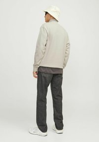 Vyras, dėvintis smėlio spalvos kibiro formos skrybėlę, šviesiai gelsvą džemperį, tamsiai pilkas džinsines kelnes ir baltus sportbačius, stovintis nugara į žiūrovą prieš paprastą foną.